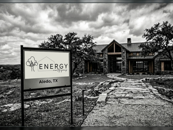 A moody, desaturated black-and-white photograph of a luxurious, rustic-modern ranch-style home in the Aledo, Texas landscape under a dramatic, cloudy sky. In the foreground, a prominent real estate yard sign with a black frame displays the 'Energy Realty Group' logo, which features simple line-art icons of a house and trees above the company name. Text at the bottom of the sign confirms 'Aledo, TX'. The sign is positioned next to a rustic, stone-paved path that leads directly to the house's inviting front entrance. Large oak trees frame the stone structure of the house, which features extensive wood trim and many windows. A dark border frame surrounds the entire image.