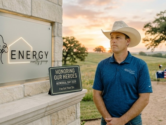 Respectful sunrise portrait of Marine Veteran and Real Estate Broker Nick Getzendanner in a cowboy hat leaning against a stone wall. He is looking off-camera. Behind him, the backlit ENERGY Realty Group logo (with tree/house line-art) from image_0.png is visible, along with a integrated digital display showing 'HONORING OUR HEROES' and 'Memorial Day 2026'. The background shows a gentle North Texas landscape in Parker or Tarrant County.