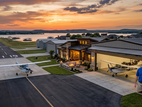 Aerial view of a custom hangar home at dusk in a Granbury TX aviation community near the lake, featuring a private plane on the asphalt taxiway next to the runway.