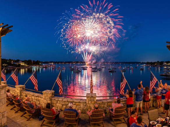 A high-angle view of a festive crowd gathered on a stone patio with string lights and American flags, watching a vibrant red, white, and blue fireworks show explode over Lake Granbury, Texas, during the 250th Independence Day celebration.